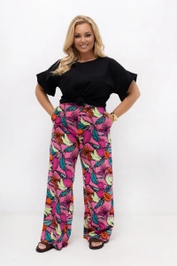 Spodnie damskie plus size Marisa w różowy wzór owocowy z szerokimi nogawkami. Model prezentuje spodnie z wysokim stanem, elastyczną gumką w pasie i kieszeniami, zestawione z czarną bluzką.