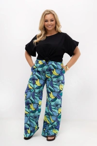 Spodnie damskie plus size Marisa w turkusowym kolorze z egzotycznym owocowym wzorem. Model prezentuje spodnie z wysokim stanem, elastyczną gumką w pasie, kieszeniami oraz szerokimi nogawkami.