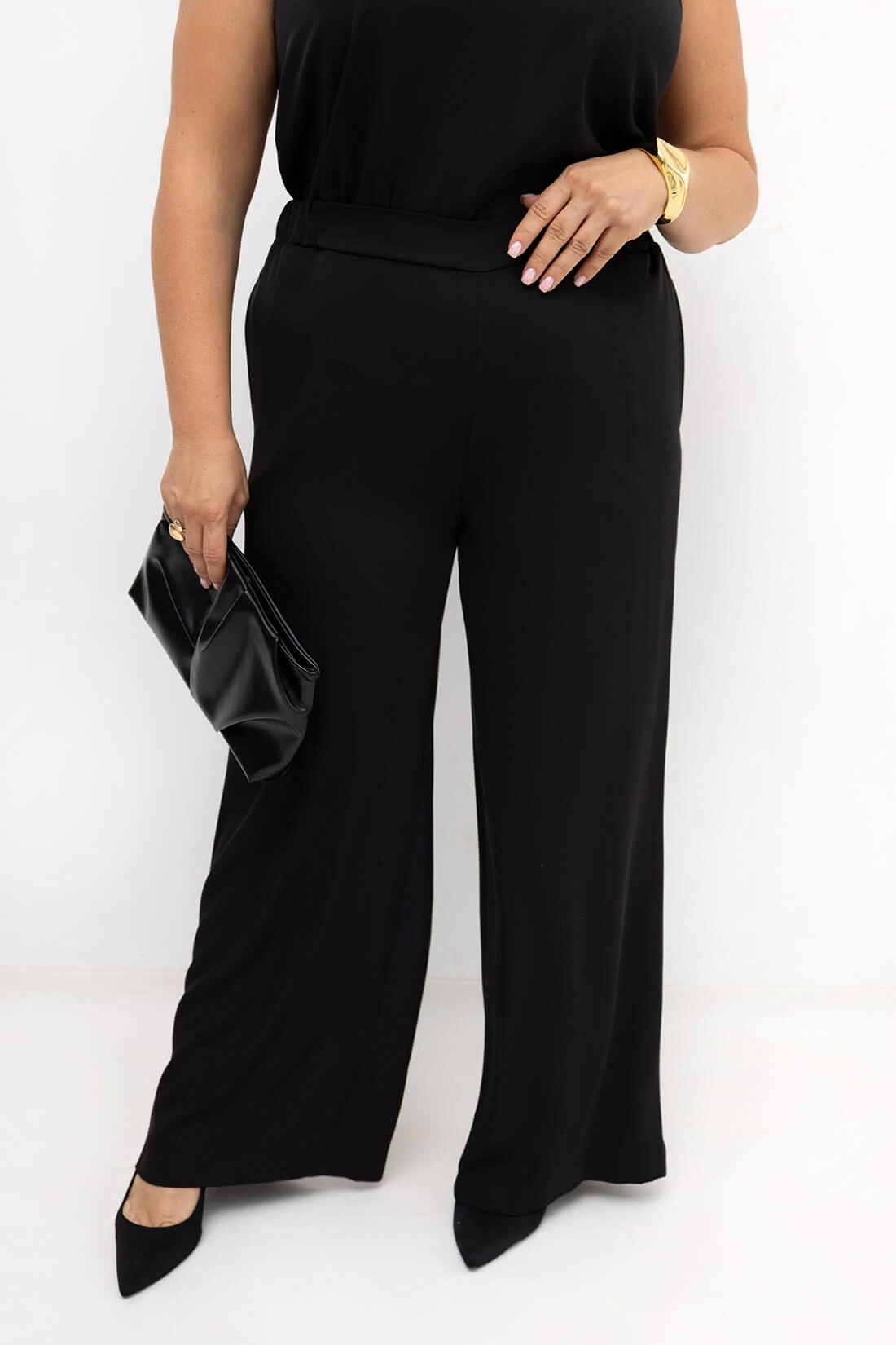 Czarne eleganckie spodnie damskie PLUS SIZE Elisa – przód stylizacji z szerokimi prostymi nogawkami. Ujęcie prezentuje dopasowanie spodni oraz przód czarnego topu z dekoltem w kształcie litery V.